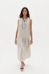 Vestido midi de lino color beige, sin mangas, con cuello redondo y detalles de ondas en las sisas. Presenta un corte con volante fruncido en la parte inferior.