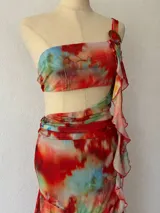 Vestido corto de dos piezas (crop top y minifalda) con estampado abstracto en tonos rojos, naranjas, amarillos y azules. El top es tipo bandeau con un tirante que incorpora un volante drapeado que cae sobre el abdomen. La minifalda es ajustada con volante en el ruedo.