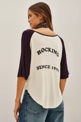 Remera estilo beisbolera con mangas raglán en color violeta oscuro y cuerpo en tono crema. Presenta una estampa frontal con texto en color negro.