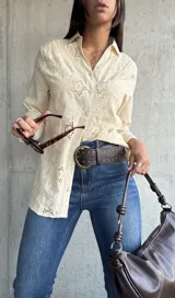 Camisa de color beige con diseño de broderie anglaise, cuello clásico, cierre frontal con botones y mangas largas.