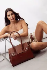 Cartera de mano estructurada de cuero vacuno rojo, con doble asa superior y detalles de charol en la parte superior. Cuenta con cierres metálicos y apliques decorativos laterales con hebillas.