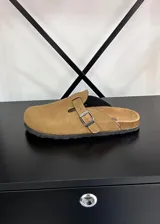 Zuecos de gamuza color beige, con diseño cerrado en la parte delantera, tira ajustable con hebilla metálica y suela de goma con borde de corcho.