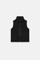 Chaleco negro de cuello subido y manga sisa, con cierre frontal de cremallera metálica. Presenta detalles de lazadas ajustables en el cuello y la cintura.