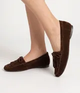 Mocasines de mujer Satinato confeccionados en cuero de gamuza marrón, con flecos, moño decorativo y costuras visibles en la capellada. Presentan punta redondeada y suela baja.