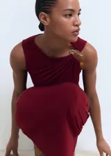 Vestido midi entallado de color bordó, sin mangas, con cuello redondo y detalle drapeado en el lateral de la cintura. Presenta una joya decorativa en uno de los hombros.