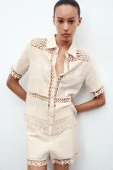 Blusa de manga corta confeccionada en algodón, con cuello clásico y cierre frontal de botones. Presenta un diseño combinado con paneles de tejido bordado floral y detalles de crochet en cuello, mangas, frente y ruedo.