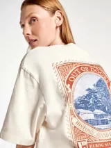 Camiseta de corte oversized color beige, con cuello redondo y mangas cortas. Presenta un estampado frontal con diseño de palmeras y texto en tonos azul y bordó con efecto desgastado.