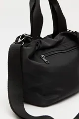 Cartera tipo bolso de nylon negro, con diseño utilitario y formato flexible que se ajusta con cordón en la parte superior. Posee asas de mano y una correa ajustable y desmontable para llevar cruzada. Incluye bolsillo frontal con cierre y herrajes metálicos.