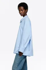 Camisa oversize de algodón color celeste, con cuello camisero, botones frontales y mangas largas ranglán con puños abotonados. Presenta pespuntes a contraste en toda la prenda.