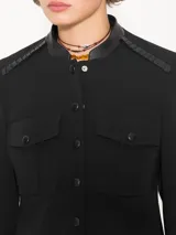 Chaqueta negra con cuello mao y charreteras de cuero ovino tableado en los hombros. Tiene acceso mediante broches forrados de cuero, bolsillos delanteros plaqué con tapa y fuelle, y godet en la espalda baja.