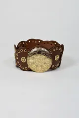 Cinto ancho de cuero marrón con diseño de piezas circulares unidas, tachas doradas y una gran hebilla metálica circular con grabado ornamental.