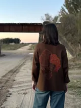 Camisa marrón de lana con cuello camisero, cierre frontal con botones y bolsillos de parche con apliques de tejido artesanal. Manga larga con puños abotonados y corte holgado.