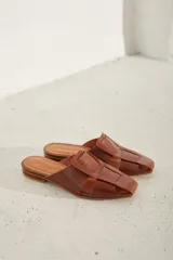 Zueco plano de cuero color dorado, con diseño destalonado, puntera cuadrada y tiras anchas entrelazadas en el empeine.