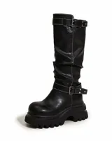 Bota bucanera de caña alta con diseño arrugado, confeccionada en cuero sintético negro con costuras blancas a la vista. Presenta dos correas ajustables con hebillas metálicas, una en la parte superior de la caña y otra en el tobillo, y una suela chunky de goma con plataforma dentada.