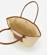 Bolso de playa tipo tote bag, tejido en fibra de papel color natural, con bordes y detalles en cuero sintético marrón. Posee asas de hombro en cuero sintético marrón y un parche cuadrado con logo grabado en el frente.