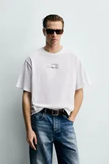 Camiseta de corte boxy fit, color blanco, con cuello redondo y manga corta. Presenta una aplicación de etiquetas decorativas a contraste en el centro del pecho.