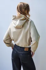 Cazadora color beige con capucha ajustable, manga larga con puño elástico, bolsillos frontales con solapa, bajo ajustable y cierre frontal con cremallera y botones a presión.