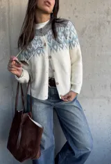 Cardigan de tejido de punto con diseño jacquard en la parte superior, cuello redondo y cierre frontal con botones. Presenta un patrón geométrico en tonos gris y azul sobre base color crema.