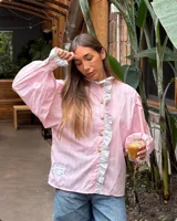 Camisa de manga larga abullonada en color rosa con rayas verticales finas. Presenta un diseño con volados en el cuello y a lo largo de la botonadura frontal, puños con detalle de rayas contrastantes y botones decorativos dorados.