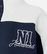 Buzo tipo polo de textura waffle con diseño color block en blanco y azul marino. Presenta cuello alto con cierre de botones, mangas largas y bordados decorativos en el frente.