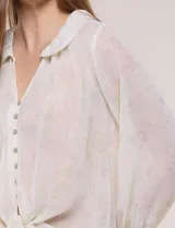 Blusa confeccionada en gasa semitransparente con sutil acabado plisado y estampado floral en tonos suaves. Presenta cuello solapa redondeado, escote en V y botonadura frontal. Mangas largas con puños elásticos y ruedo anudado en la parte inferior frontal.