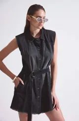 Vestido camisero negro de denim rígido liviano sin mangas, con acceso por botones en delantero y cinto con hebilla metálica plateada.