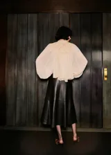 Blusa de color crema con diseño evasé, cuello redondo y mangas largas abullonadas. Presenta un panel superior de encaje detallado y un cierre de botón en la parte posterior del cuello.