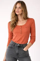 Remera blanca de manga larga, entallada, con cuello redondo y una hilera de botones en el frente. Incluye un pequeño bolsillo decorativo en el pecho.