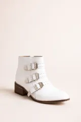 Botineta blanca de cuero, con taco cuadrado de madera, punta afilada y tres tiras con hebillas metálicas y tachas decorativas.