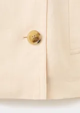 Chaqueta crop de lino color beige, diseño recto, cuello alto tipo embudo, manga larga, dos bolsillos laterales y cierre cruzado de doble botonadura metálica dorada con detalles en los hombros.