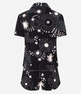 Conjunto de pijama de dos piezas confeccionado en viscolycra, compuesto por una blusa de manga corta con cuello solapa y botones frontales, y un short con cintura elástica y lazo. Estampado integral de temática espacial con estrellas, planetas y explosiones en tonos rosados y blancos sobre fondo negro.