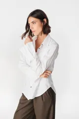 Camisa blanca de algodón con cuello clásico, cierre frontal de botones y puños anchos desmontables. Presenta un diseño con tablas decorativas y terminación redondeada en los laterales.