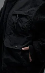Campera estilo bomber unisex, confeccionada en una combinación de corderito sintético y paneles de ripstop. Presenta un diseño utilitario con múltiples bolsillos frontales con cierres y broches a presión, cuello, puños y ruedo en ribb elástico.