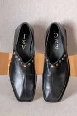 Zapato de cuero vacuno color negro, con punta cuadrada y detalle de tachas metálicas planas en el borde superior. Presenta costura central en el empeine y taco geométrico.