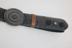 Cinturón de cuero color gris con efecto desgastado, hebilla metálica grande y redonda con diseño grabado, pasador marrón y tachas decorativas.