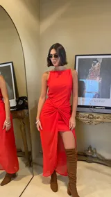 Vestido largo rojo con diseño asimétrico, escote redondo y detalle de nudo drapeado en la cintura que crea una abertura frontal, dejando ver una falda corta interior.