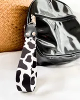 Llavero de muñeca con cinta de tela estampada con diseño de vaca en blanco y negro. Tiene un conector de cuero sintético negro con remaches plateados y un gancho metálico plateado.