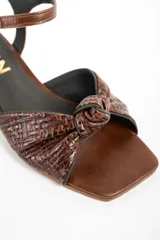 Sandalias de cuero marrón con taco cuadrado medio, tira en el tobillo con hebilla ajustable y diseño trenzado con nudo en la parte delantera.