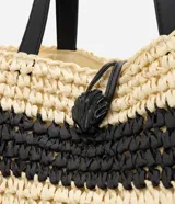 Cartera tipo saco shopper confeccionada en paja trenzada, con diseño de rayas en color natural y negro. Cuenta con asas de hombro de material sintético y cierre con cordón ajustable con detalle de concha.