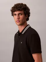 Chomba de piqué Calvin Klein color negro, con cuello clásico y ribetes a rayas blancas, cartera de dos botones y pequeño monograma CK bordado en el pecho.