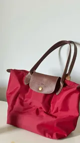 Bolso tipo shopper de cuerpo plegable en color crema o blanco roto, con asas y solapa de cierre en cuero marrón texturizado. Presenta un bolsillo exterior frontal y cierre principal con broche metálico.