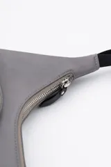 Cartera bandolera color gris de material sintético con correa ajustable negra y hebilla. Posee dos bolsillos frontales exteriores, uno con cierre y tirador largo y otro con solapa y hebilla decorativa.