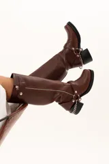 Bota de caña alta estilo biker confeccionada en cuero color chocolate. Presenta un cierre metálico lateral que recorre toda la caña, taco cuadrado y plataforma. Incluye un sistema de estribo y cadena metálica desmontable en la zona del tobillo, permitiendo diversas formas de uso.