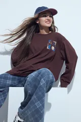 Remera de manga larga color marrón chocolate, con corte holgado y cuello redondo. Presenta un estampado pequeño en el pecho con motivos de lunas y rostros en tonos azules y crema.