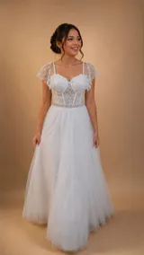 Vestido largo de novia en color blanco, con cuerpo tipo corsé de encaje con escote corazón y transparencias. Presenta breteles finos, mangas cortas caídas de encaje y una falda amplia de tul con una faja decorativa de pedrería en la cintura.