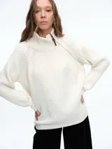 Sweater de tejido de punto acanalado en color blanco, con cuello alto y cierre metálico diagonal en el hombro. Presenta mangas largas con diseño raglán y corte holgado.