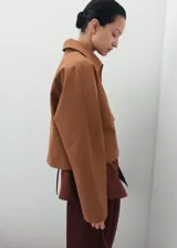 Chaqueta de corte recto color tostado, con cuello camisero, manga larga, dos bolsillos de parche con solapa en el frente y cierre frontal de botones oscuros.