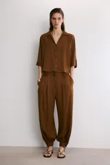 Pantalón de tiro alto en color beige, con diseño de pierna holgada y puños elásticos con cierre de botón en el bajo. Presenta cierre frontal con cremallera y doble botón en la cintura, además de bolsillos laterales.