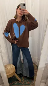 Campera tejida de punto en color marrón chocolate, con cierre frontal completo y diseño de corazón azul en el frente y en una de las mangas. Presenta detalles bordados decorativos y puños y ruedo acanalados.