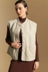 Chaleco de paño color beige, de silueta recta y diseño minimalista. Presenta cuello a la base, cierre frontal con botones grandes y bolsillos aplicados en los laterales.